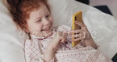 有趣的红头发小女孩与黄色智能手机躺在床上和使用智能手机。 沟通，播放，APP..